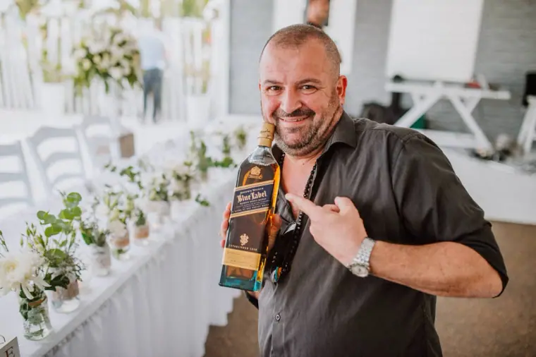 Grinning guest holding up a bottle of whisky beside a dressed reception table, Galu Seaside