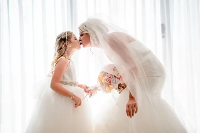 Bride bending to kiss the flower girl in a bright window