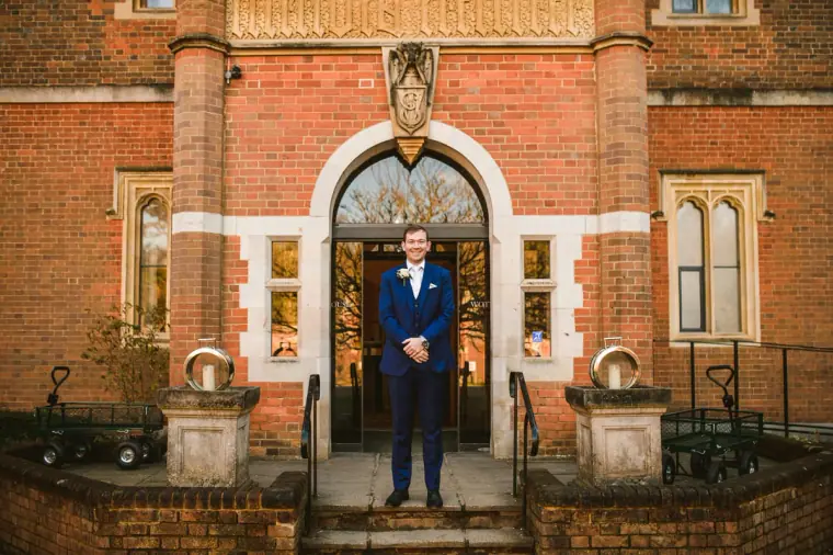 Groom in a blue suit standing in the arched red-brick entrance of Wotton House