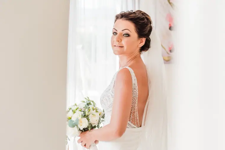 Bride glancing over her shoulder by the window, bouquet in hand