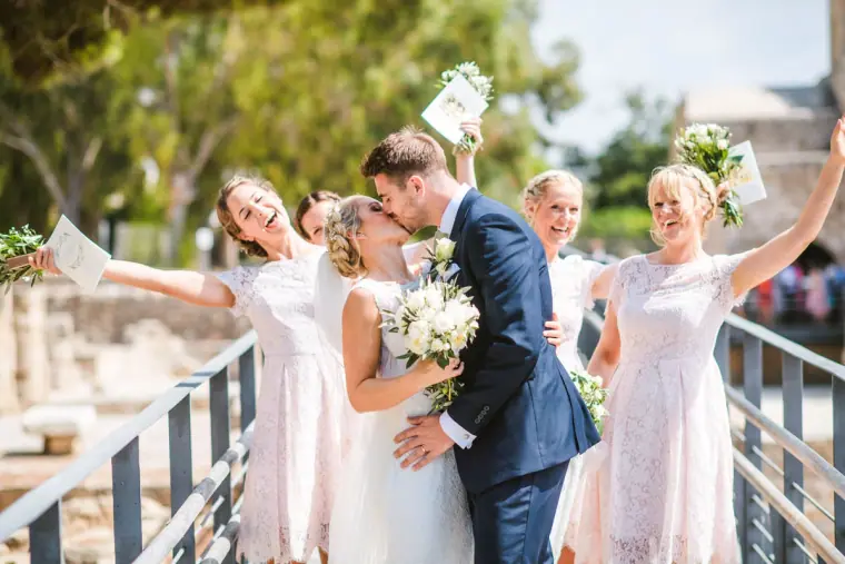 Rachel and Chris kissing as bridesmaids in blush cheer and wave behind them, Pissouri