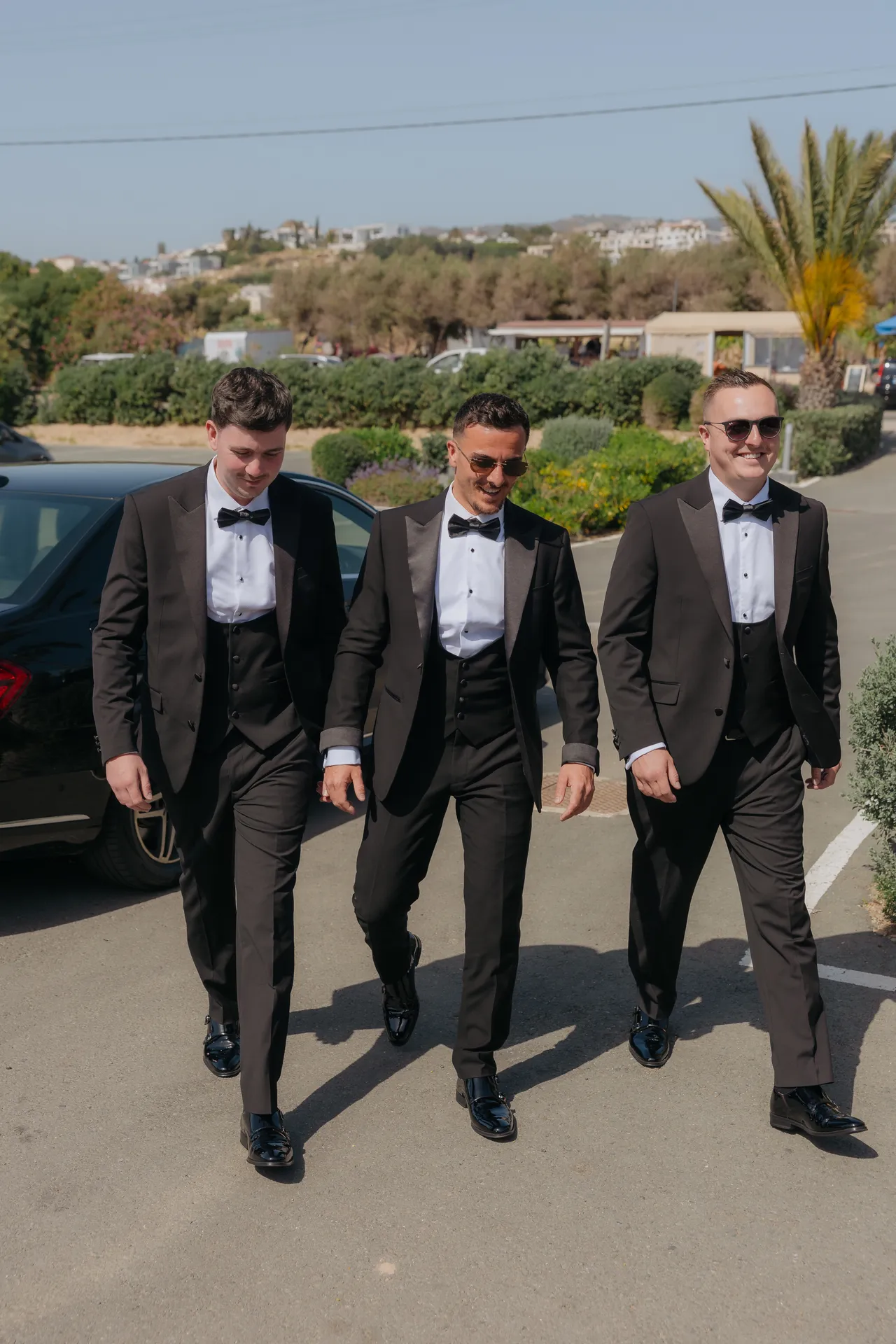 Groom and two groomsmen in tuxedos walking across the car park, sunglasses on