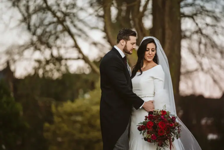 Stephanie and Jamie embracing among trees, her red bouquet held low