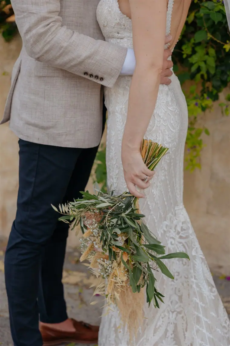 Close-up of the bride's lace gown and olive-and-pampas bouquet in the groom's embrace