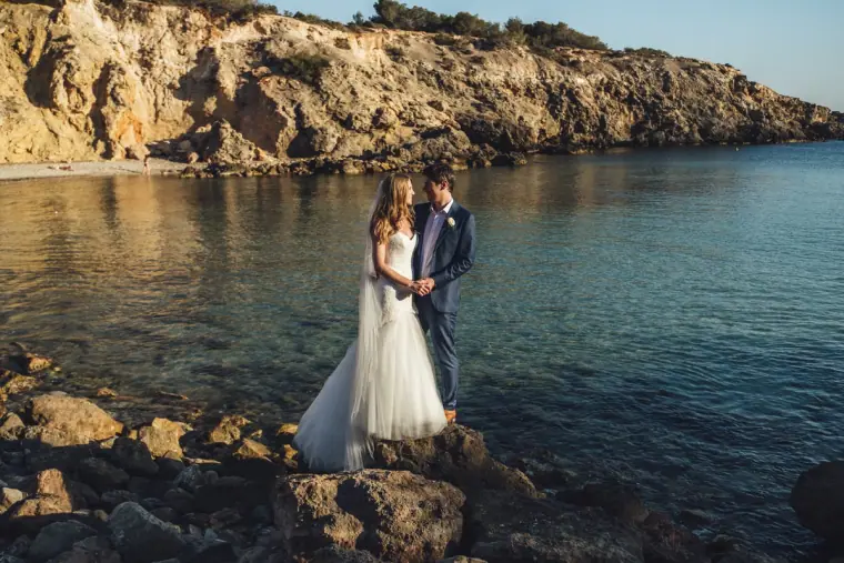 Bride and groom standing on the rocks at the water's edge in a sheltered cove