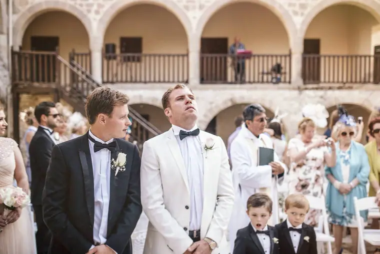 Groom and best man turning to see the bride, with guests and page boys behind