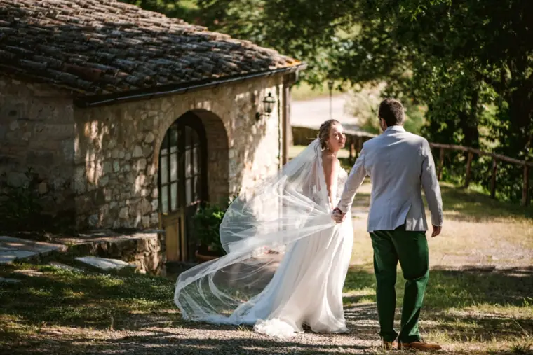 Groom leading his bride past a small stone cottage, her veil trailing on the grass