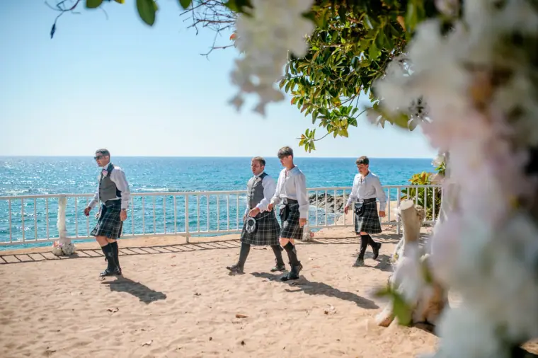 Men in kilts walking across the sandy ceremony area by the sea