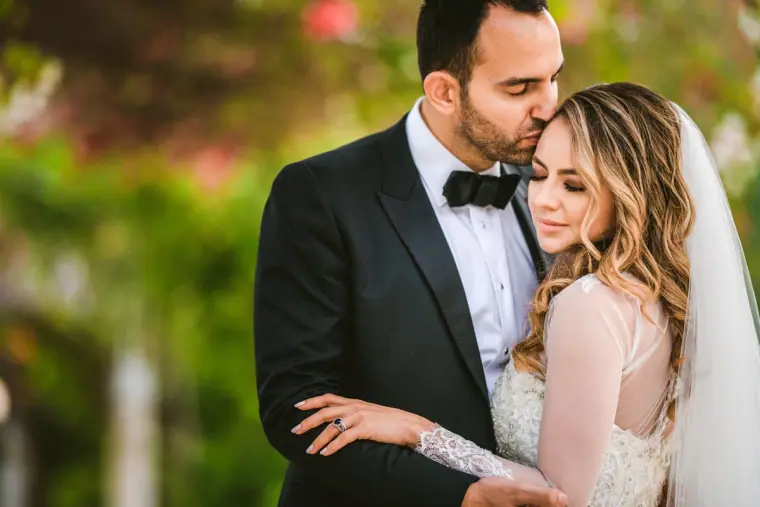 Groom kissing the bride's forehead, framed by soft pink flowers