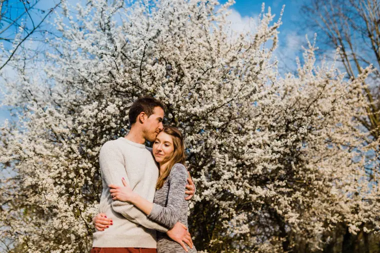 Alex kissing Jana's head in front of a tree in full white blossom