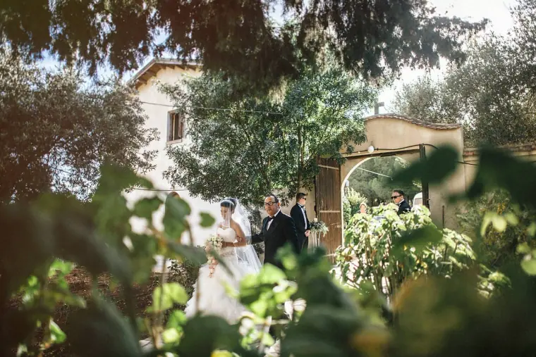 Bride walking into the courtyard through leafy trees on her father's arm