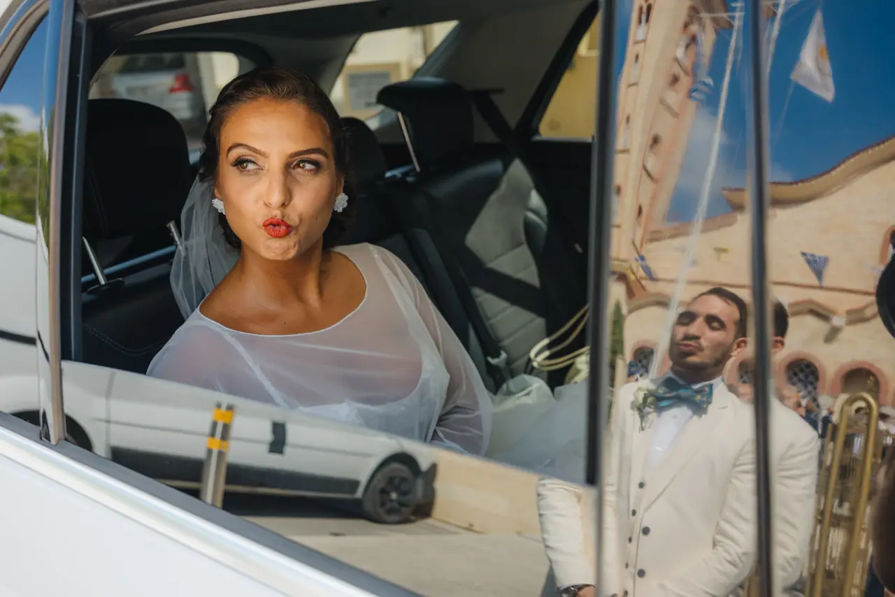 Bride blowing a kiss from a car window as the groom in a white tuxedo waits outside