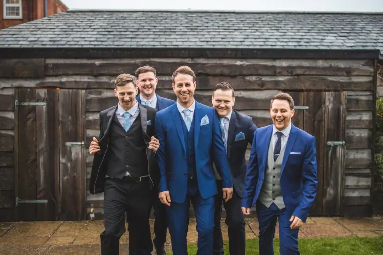 Groom and groomsmen laughing together in front of a rustic timber barn