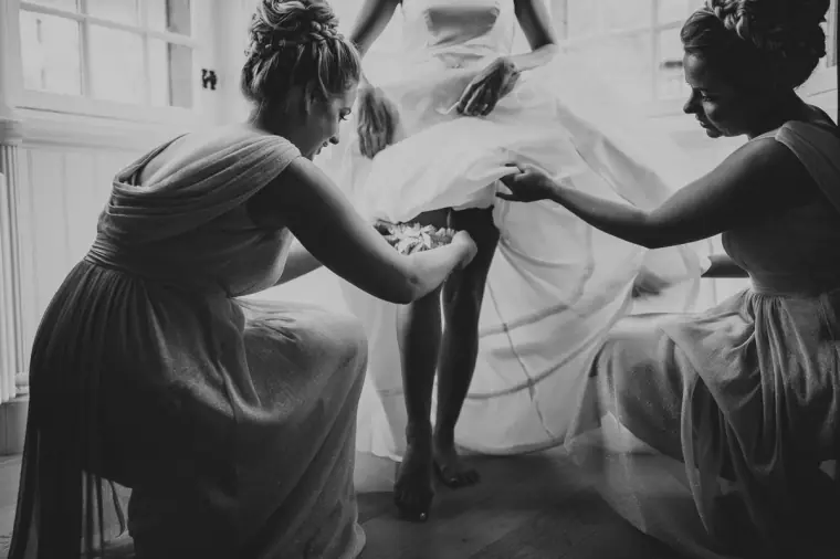 Black-and-white photo of bridesmaids lifting the bride's skirt as she steps into her gown