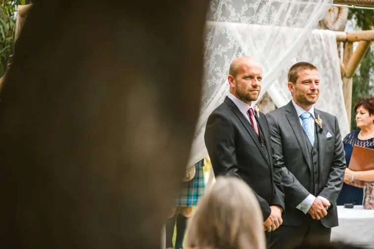 Groom and best man waiting by the lace-draped ceremony arch in the garden