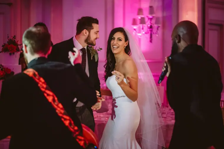 Bride and groom laughing as a live band plays in the pink-lit ballroom