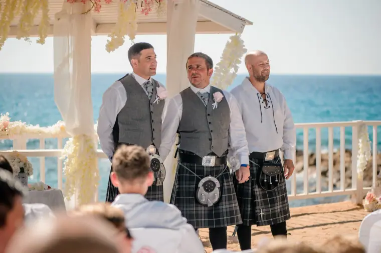 Groom in a kilt and grey waistcoat waiting with groomsmen at the seaside gazebo