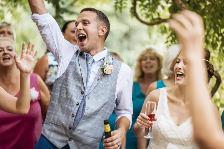 Groom cheering with a beer bottle as the bride laughs among guests