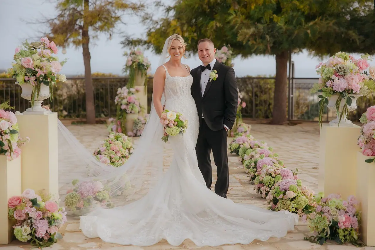 Zoe and Oliver standing together between the floral plinths at the front of the aisle, her train spread across the stone
