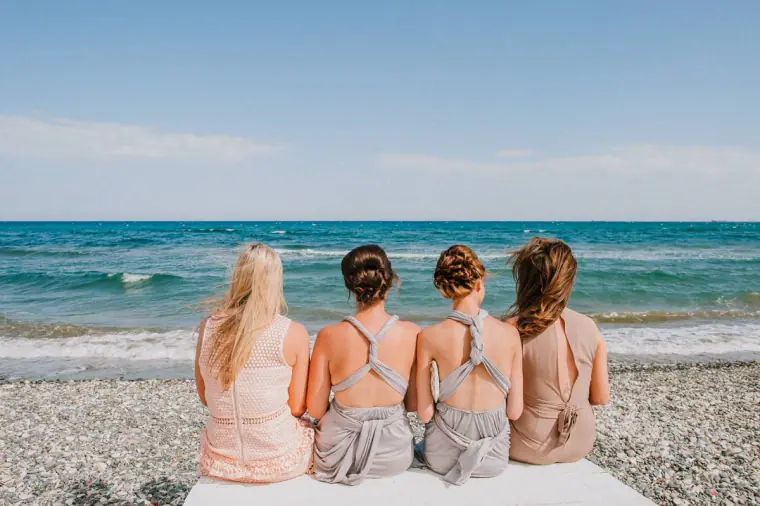 Bridesmaids in blush and grey dresses sitting together facing the sea