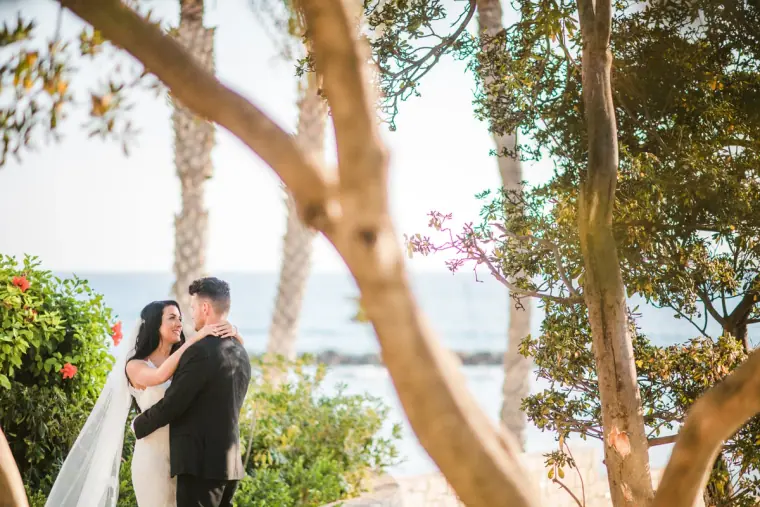 Jaclyn and Steven embracing by the sea, framed by tree branches and flowers at Almyra