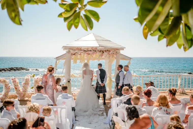 Bride and groom at the flower-draped gazebo before seated guests by the sea