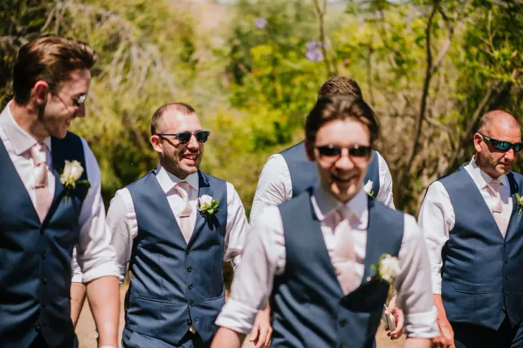 Groomsmen in sunglasses and navy waistcoats laughing as they walk together