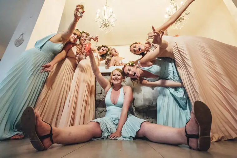 Bridesmaids in blush and pale blue gowns sprawled laughing on the floor, shot from below