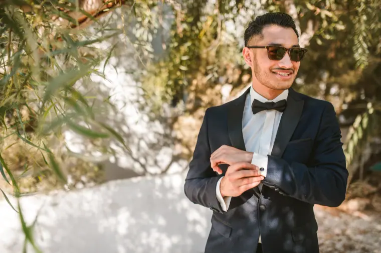 Groom in sunglasses adjusting his cufflinks beneath an olive tree