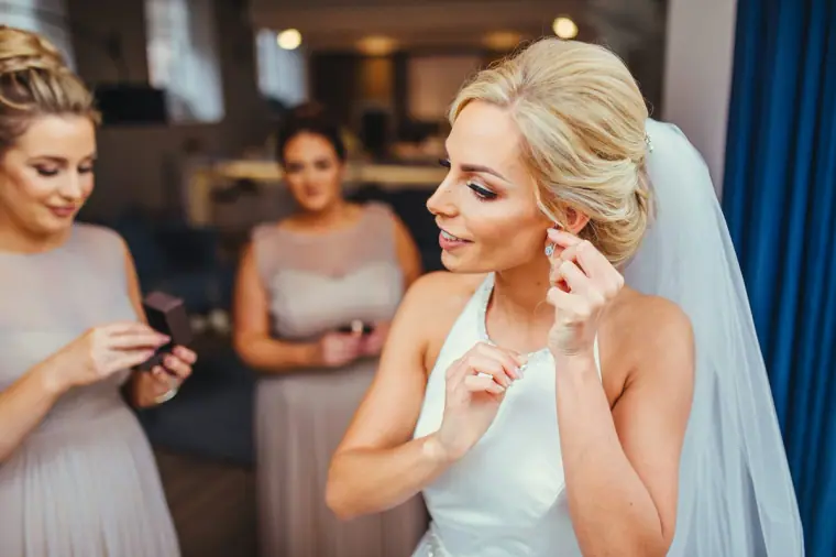 Bride fastening an earring beside bridesmaids in a softly lit room