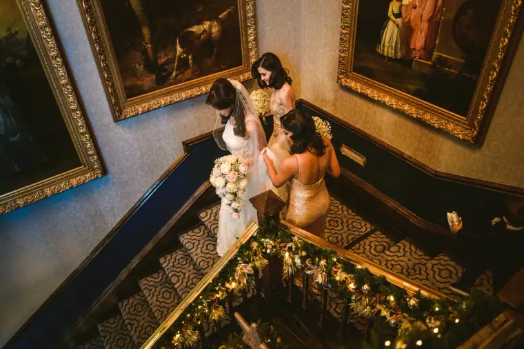 Elina and her bridesmaids descending a grand staircase beneath gilt-framed paintings