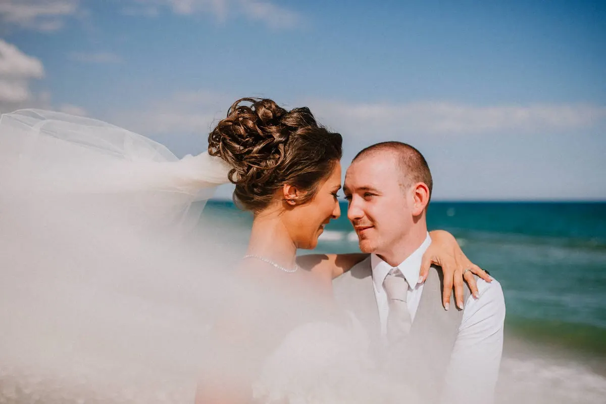 Louise and Mike touching foreheads as her veil blows in the sea breeze