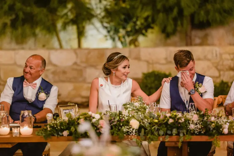 Groom wiping a tear at the top table as the bride smiles beside him