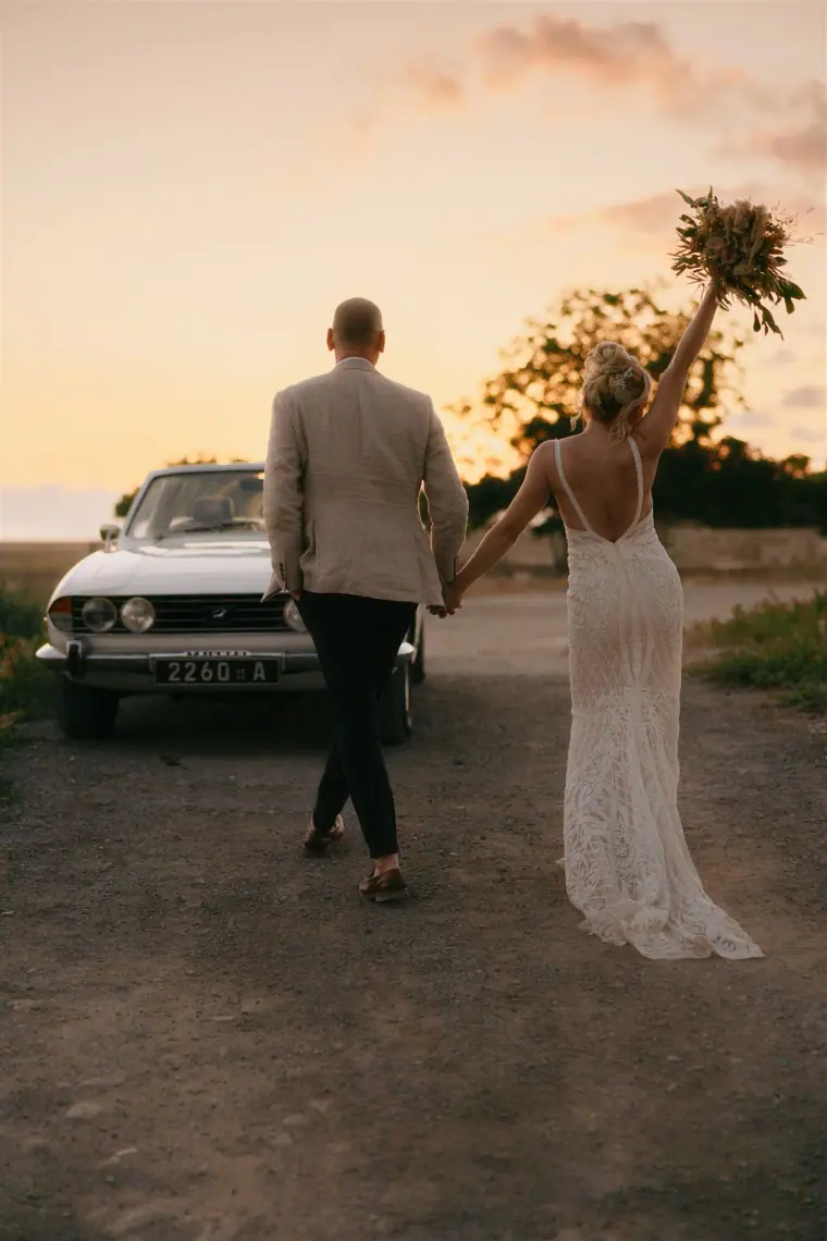 Bride raising her bouquet as the couple walk towards the vintage car
