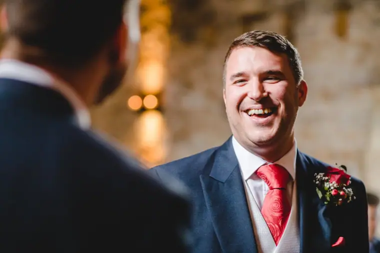 Groom in a navy suit and red tie laughing as he waits at the front