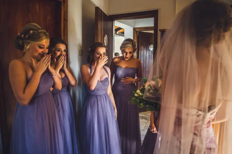 Bridesmaids in lilac covering their mouths in delight as they see the bride