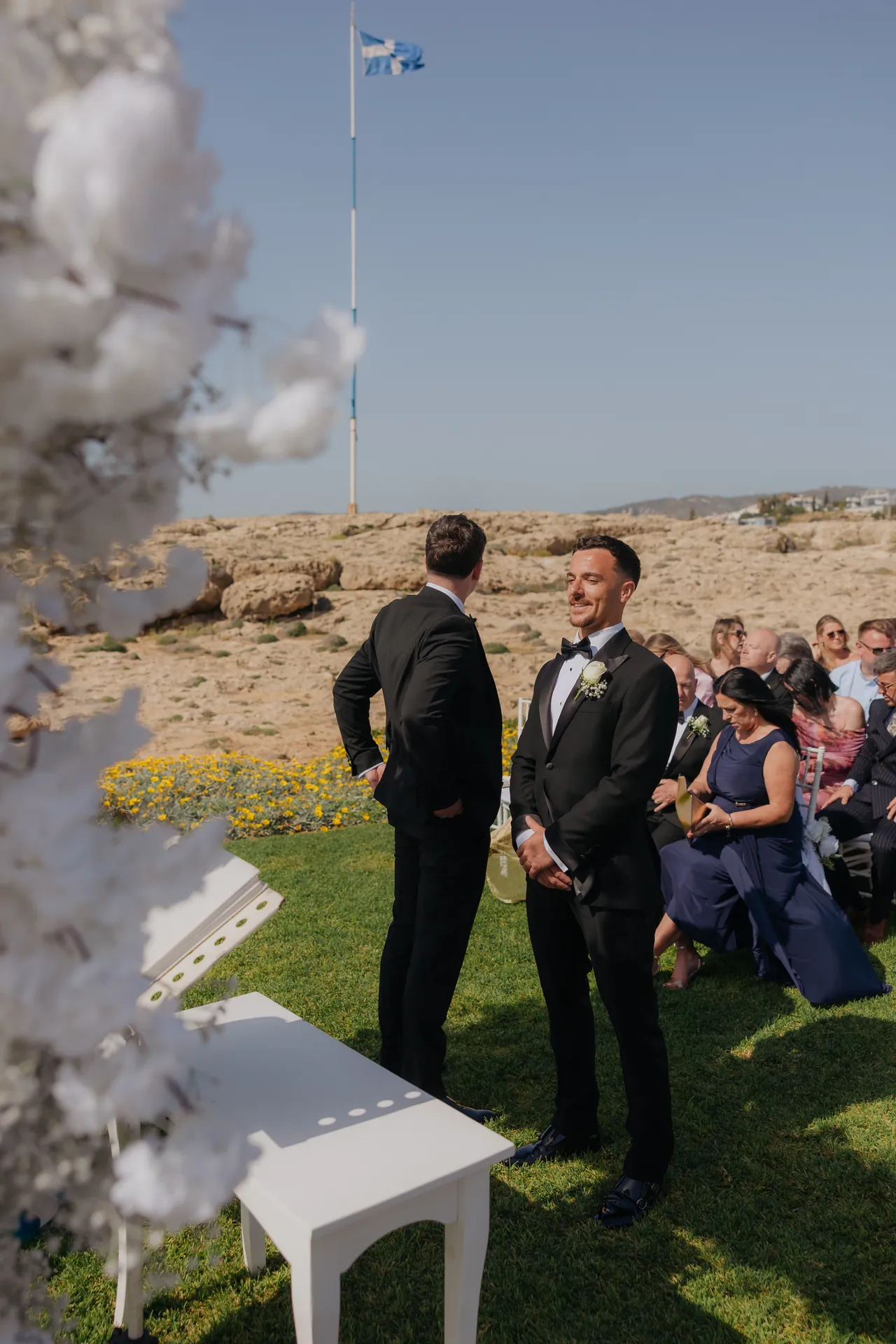 Groom waiting with his best man beside the white floral arch, guests seated behind