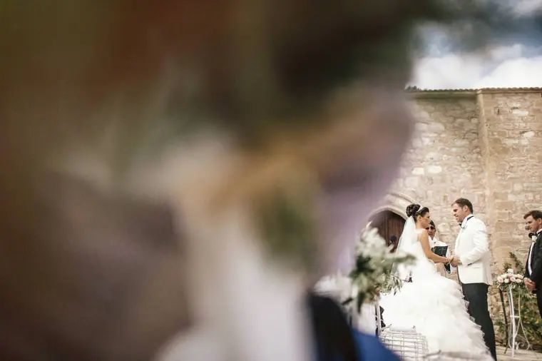 Couple at the altar glimpsed past a blurred guest, stone wall behind