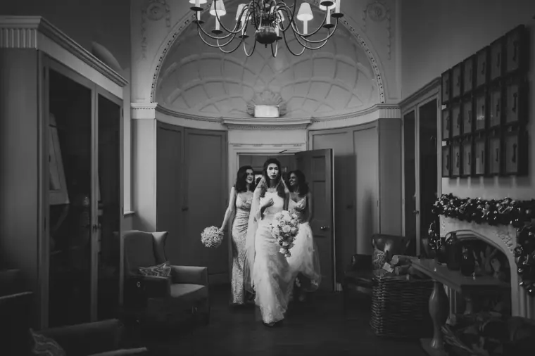Black-and-white photo of the bride arriving with her bridesmaids through a chandeliered hall