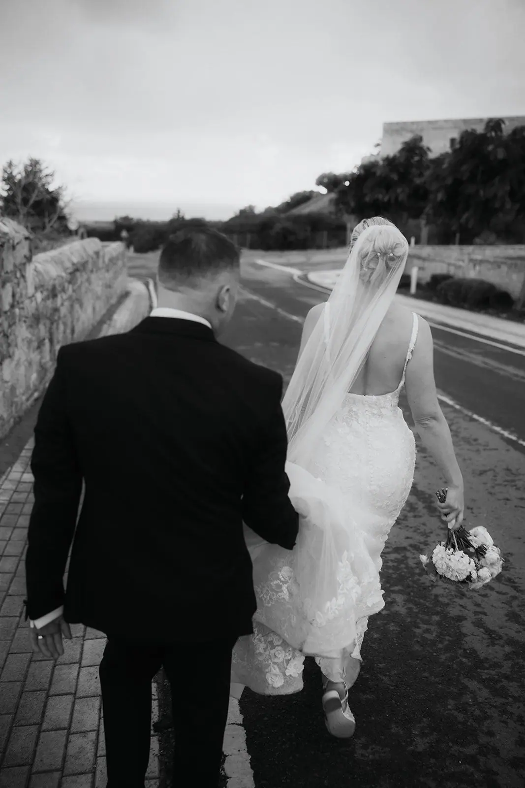 Black-and-white photo of Zoe and Oliver walking along the wall, her bouquet in hand