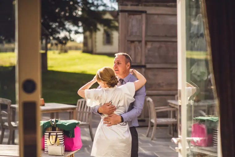 Bride in a robe hugging a man on the sunny terrace, seen through a doorway
