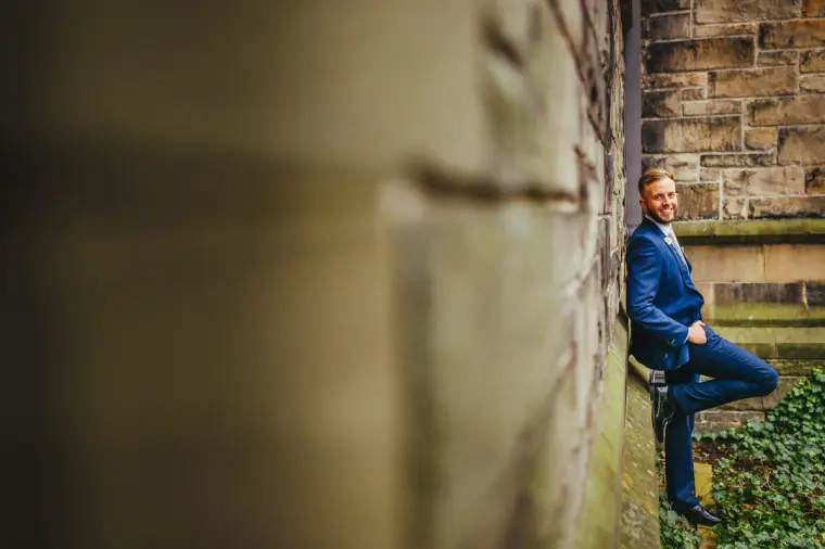 Groom leaning against a mossy stone wall, framed by a blurred pillar