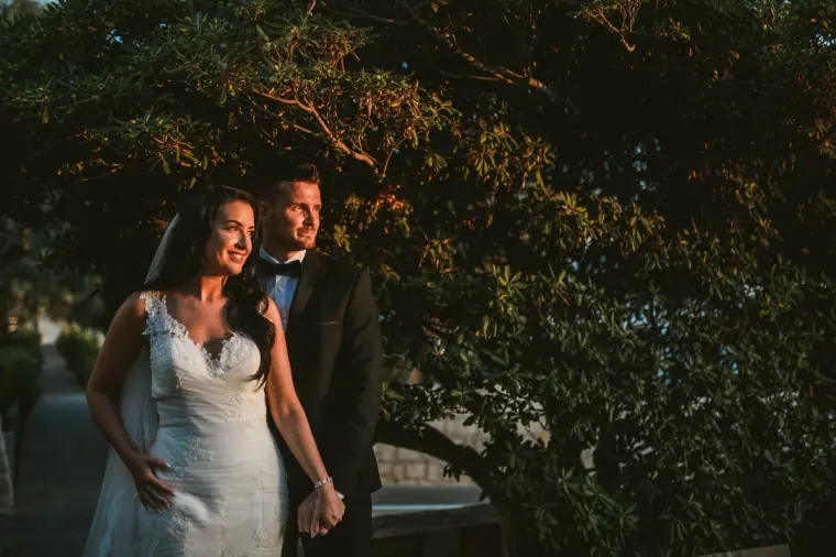 Jaclyn and Steven smiling arm in arm under dark trees at golden hour