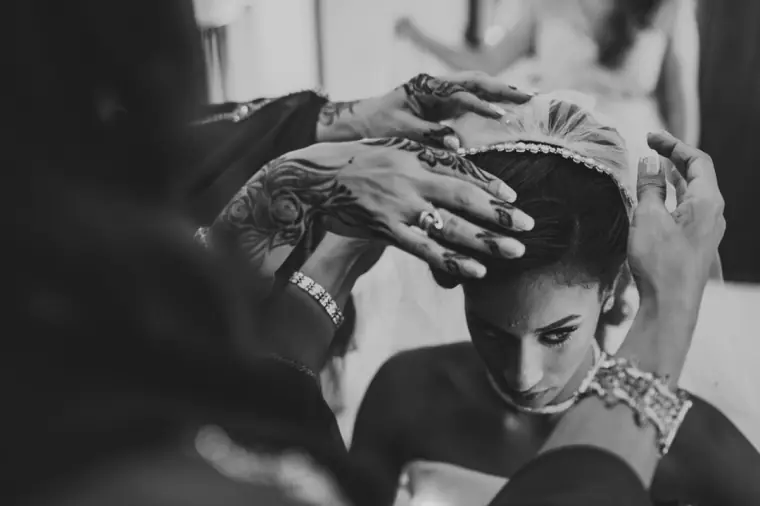 Black-and-white photo of hennaed hands placing a jewelled headpiece on the bride's hair