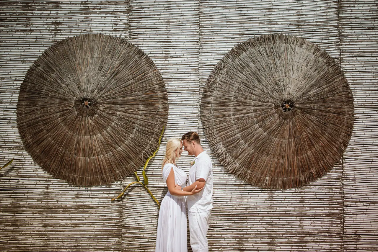 Adam and Maria touching foreheads before a woven wall hung with straw hats, Ayia Napa