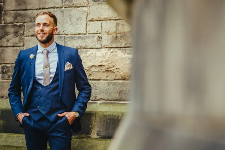 Andy standing hands in pockets in a blue suit by a sandstone wall