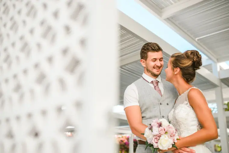 Bride and groom smiling at each other beside a white lattice wall