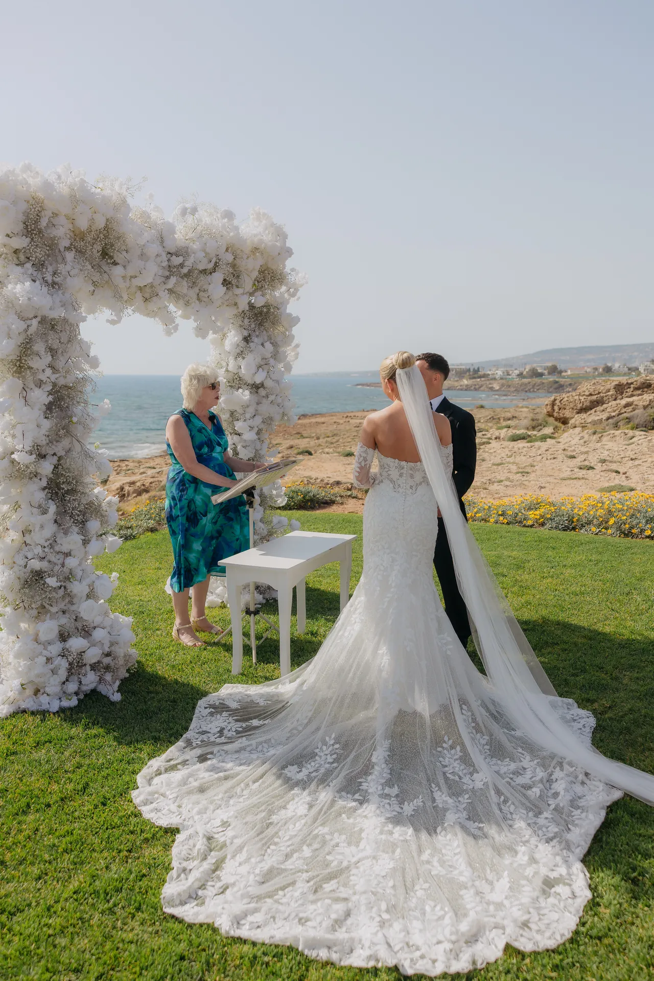 Bride with a long lace train approaching the floral arch overlooking the sea