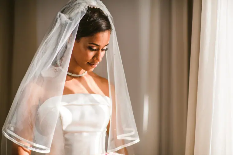Raya in her fingertip veil and strapless gown looking down by the window
