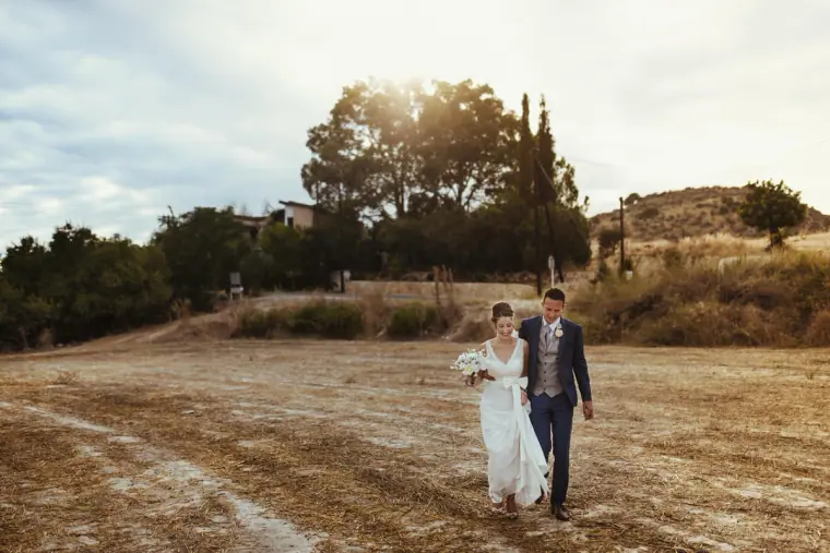 Newlyweds walking hand in hand across a dry field in the Paphos hills
